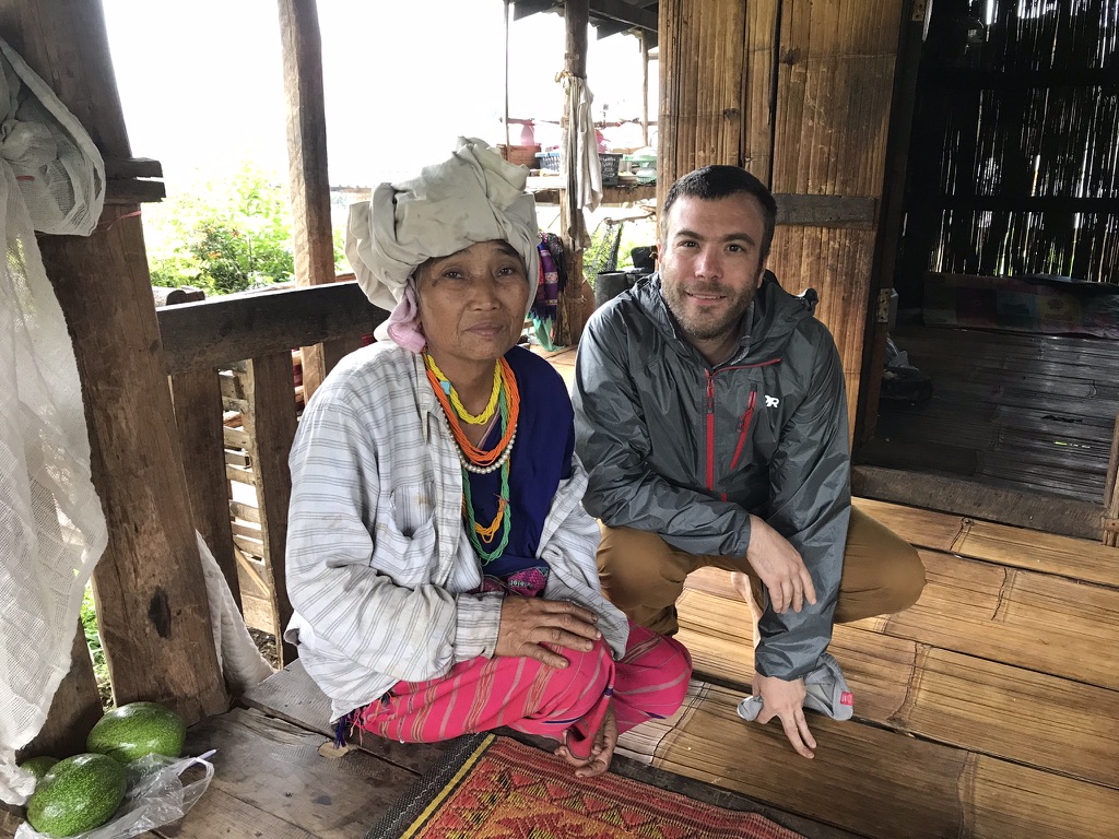 ATWR executive director Andrew Murphy crouching beside a Karen elder wearing a head wrap and beaded necklaces on the porch of her home