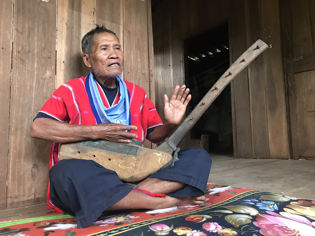 Karen elder Bookoo Sandoh sitting cross-legged on a floral mat, singing and playing the tenaku (Karen harp) in his home in Khun Tae Village, northern Thailand