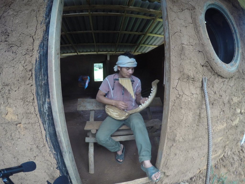 Gerse-Tu, a Karen singer, playing the tenaku (Karen harp) and panpipes in the doorway of an earthen house in northern Thailand, with ATWR recording microphones in the foreground